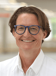 Porträt einer lächelnden Frau mit blauer Brille im weißen Kittel vor unscharfem Hintergrund.