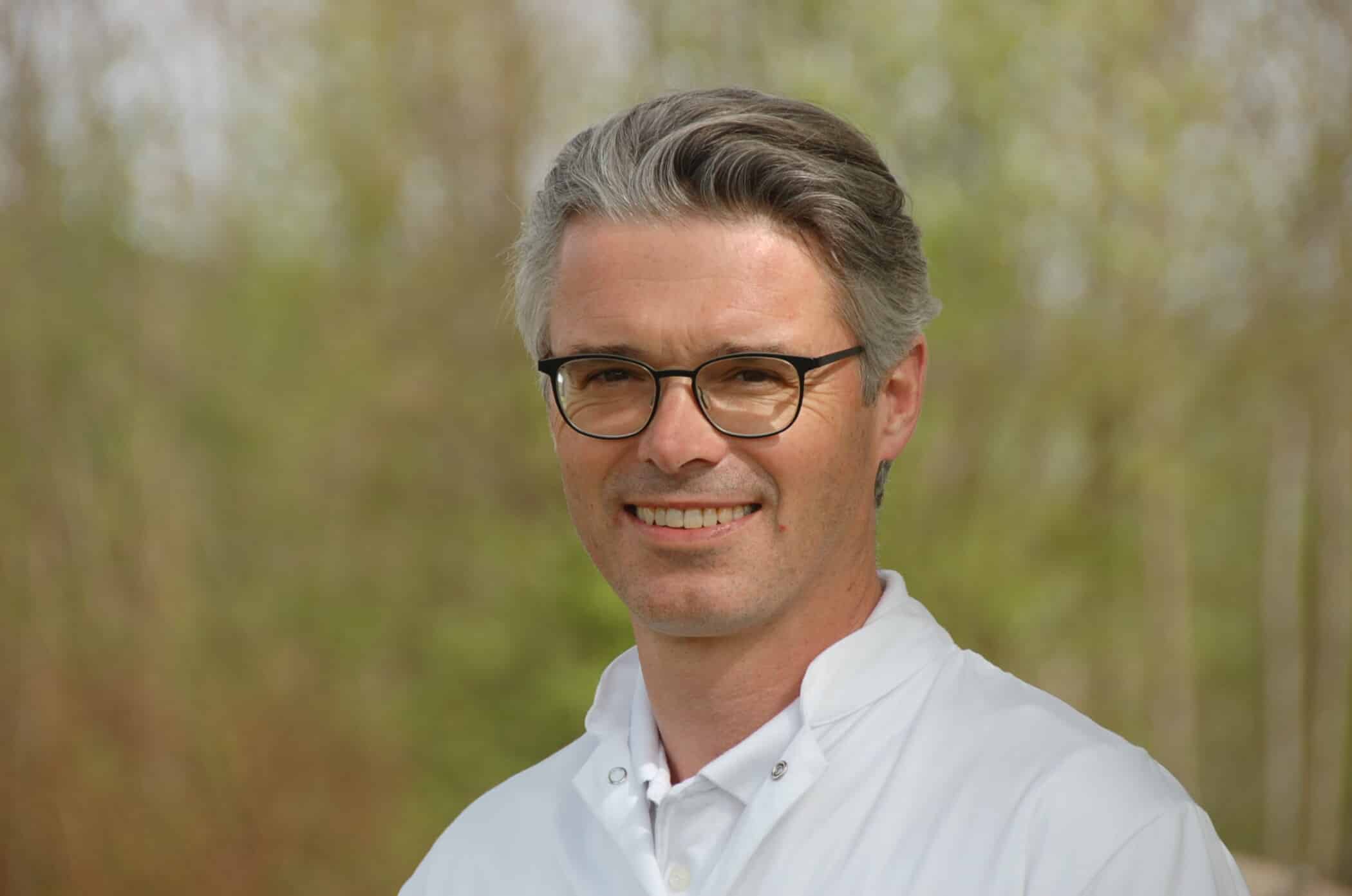 Porträt eines lächelnden Mannes mit Brille im weißen Kittel im Freien.