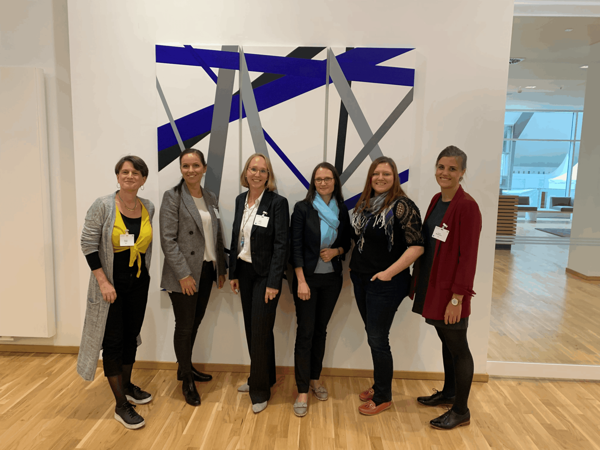 Sechs Frauen stehen lächelnd vor einem modernen Wandkunstwerk.