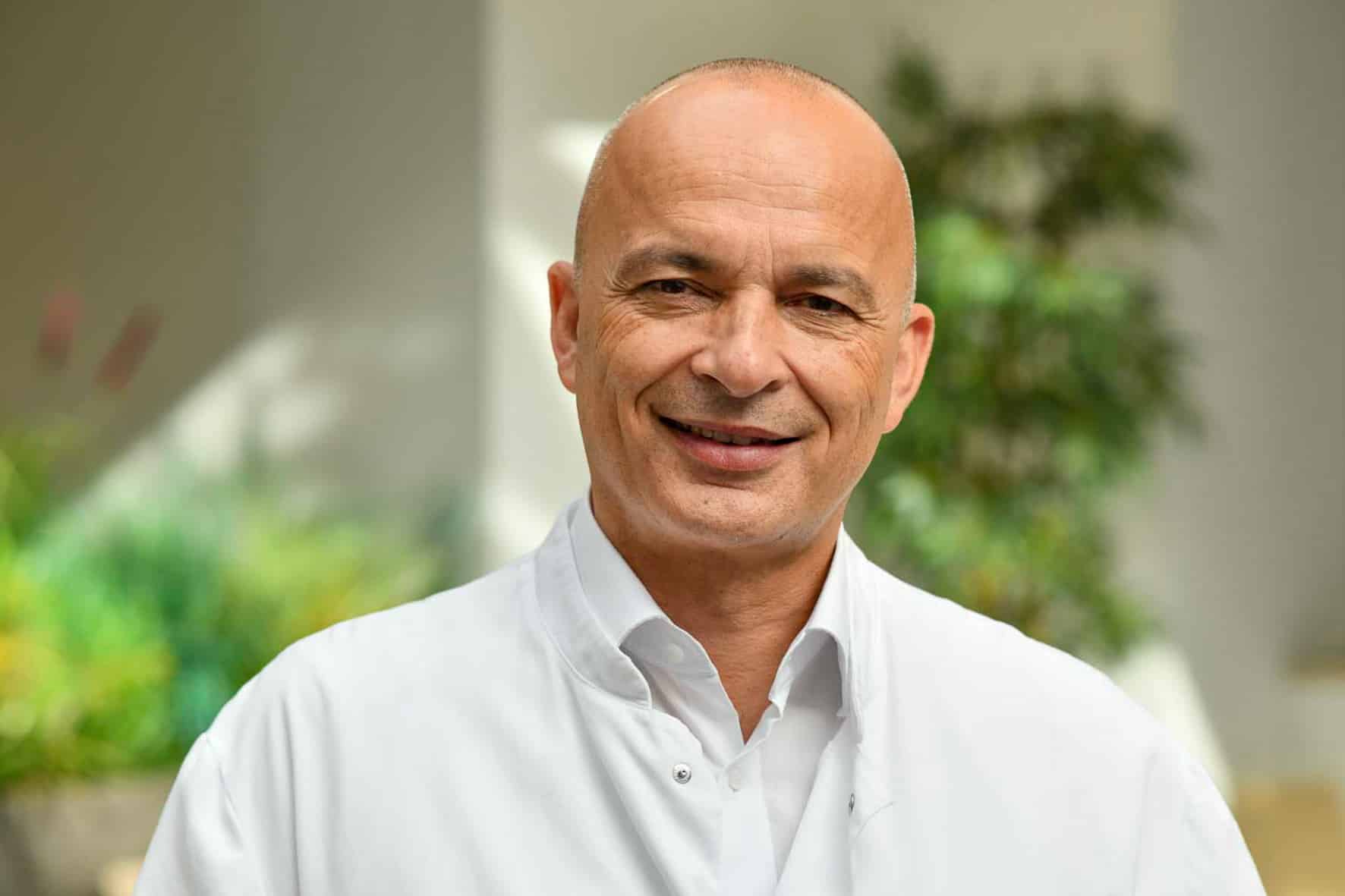 Porträt eines lächelnden Mannes in weißem Arztkittel vor unscharfem Hintergrund mit Pflanzen.