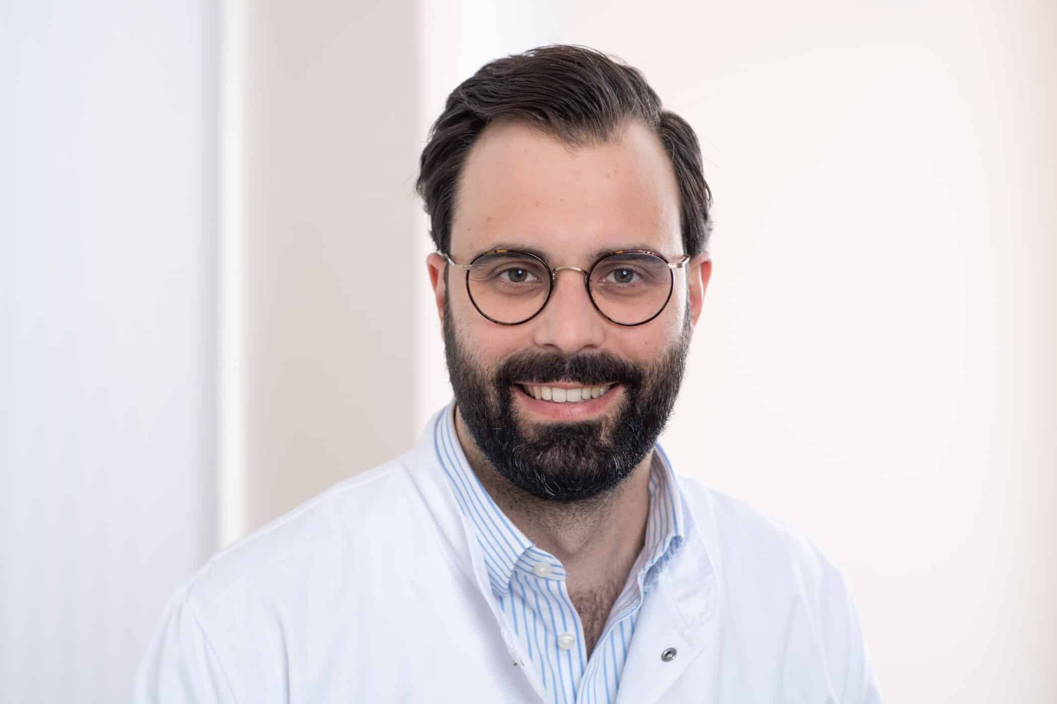 Schnorr (2) Porträt eines lächelnden Mannes mit Brille im weißen Kittel vor hellem Hintergrund.