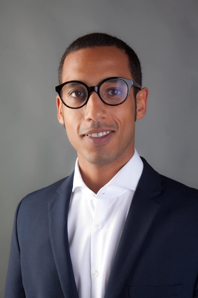 Menghesha Porträt eines Mannes mit Brille im dunklen Anzug vor grauem Hintergrund.