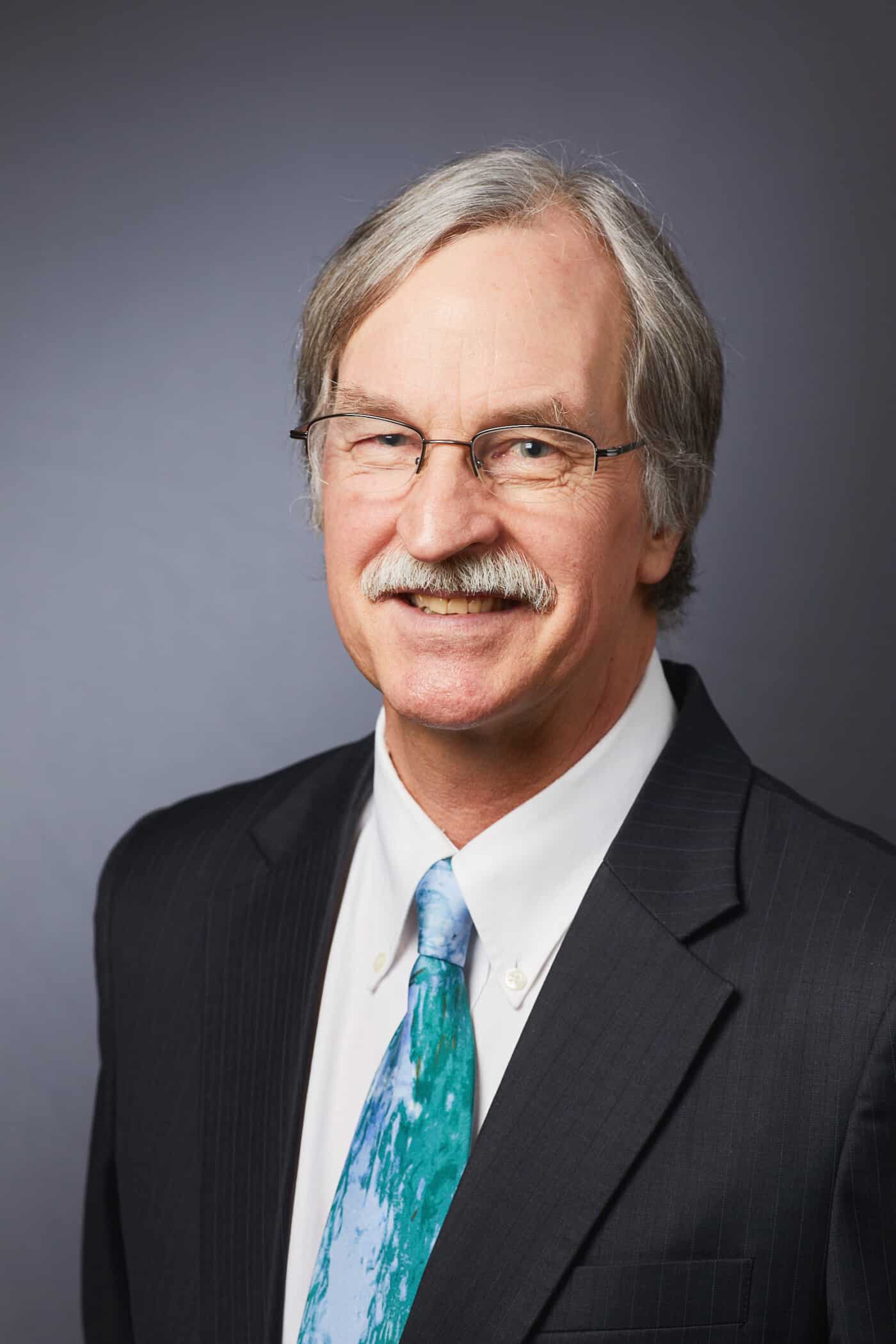 Porträt eines Mannes mit Brille und Schnurrbart im Anzug vor dunklem Hintergrund.