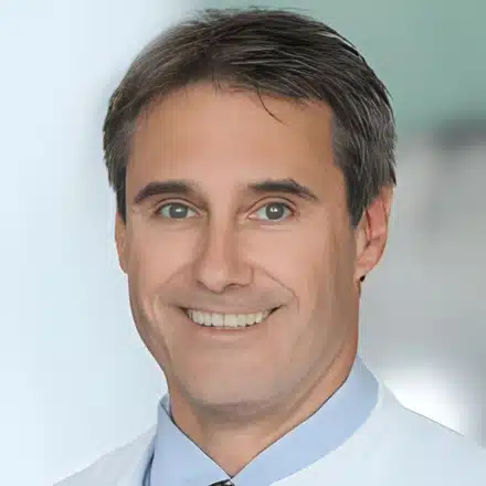 Porträt eines lächelnden Mannes im weißen Kittel vor hellem Hintergrund.